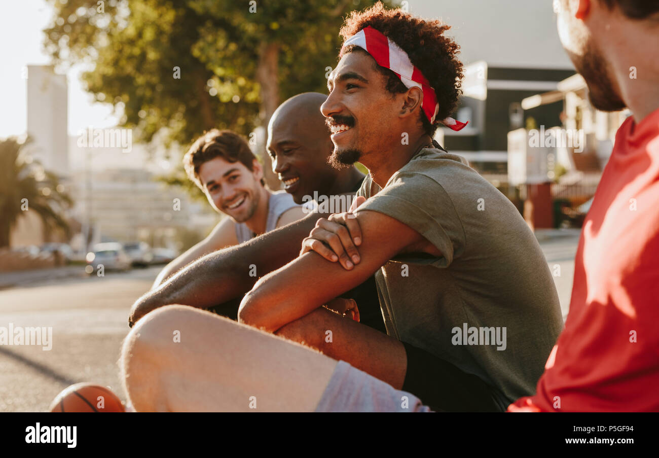 Close up of four basketball men relaxing after a game of basketball sitting on pavement. Cheerful athletes sitting on pavement and discussing. Stock Photo