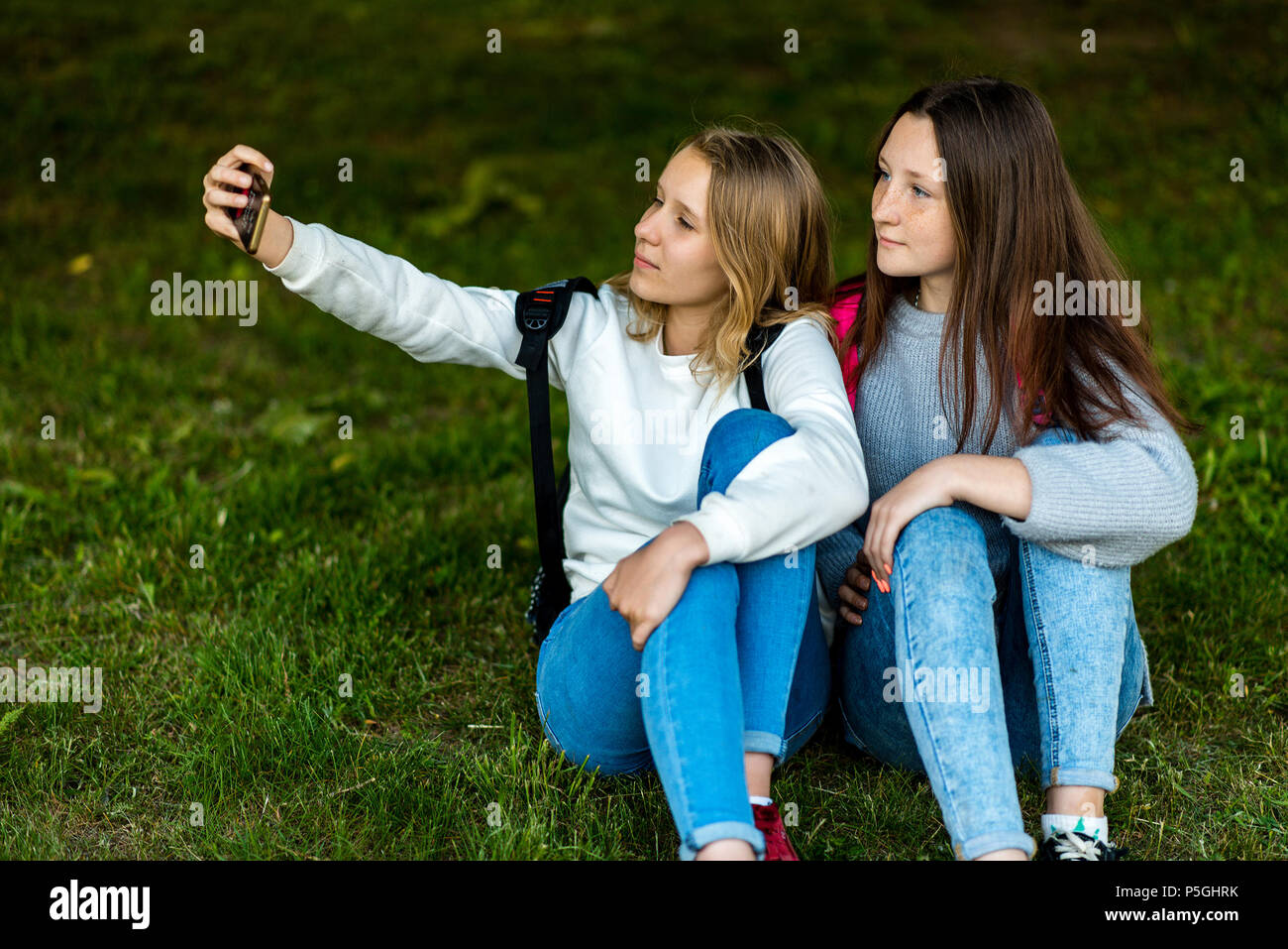 Two teenage girls. In the summer of the city park. They sit on the grass and take photos on smartphone. Behind backpacks. The concept of school friendship. Emotion pose on the phone. Stock Photo