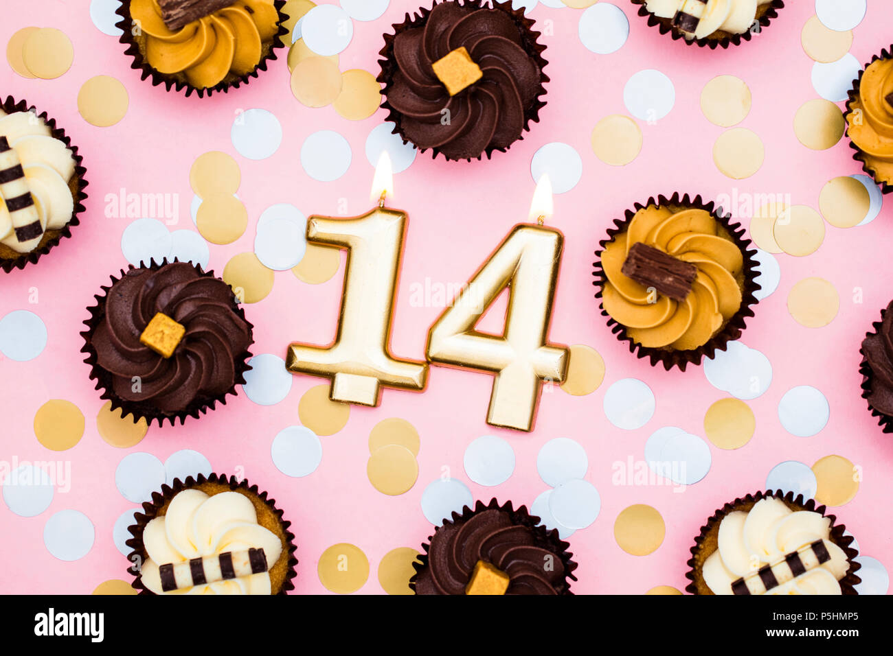 Number 14 gold candle with cupcakes against a pastel pink background Stock Photo