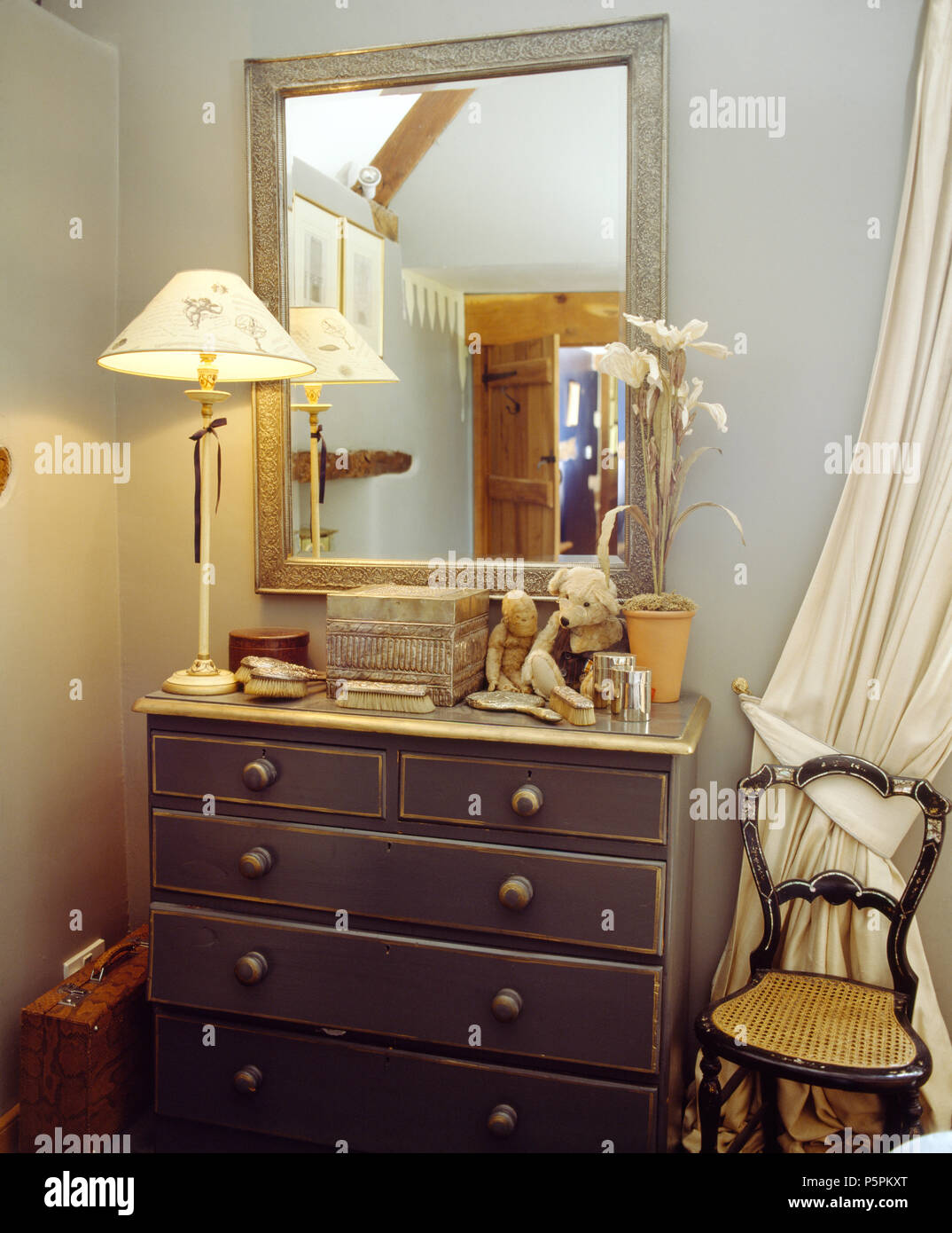 Grey painted mirror above lighted lamp on grey painted chest-of-drawers Stock Photo