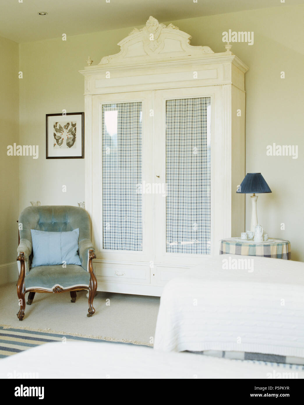 White paintedwardrobe with checked drapes behind glass doors in country bedroom with a blue velvet upholstered Victorian chair Stock Photo