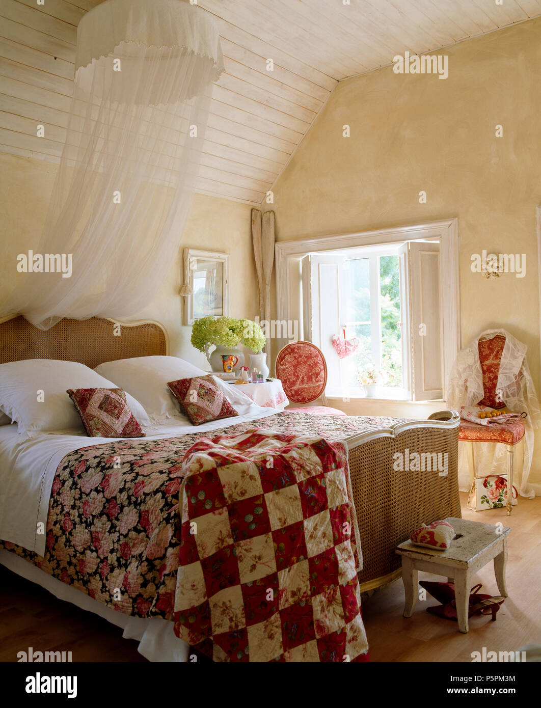 Country bedroom white voile mosquito net above antique Bergere bed with red+white patchwork and rose-patterned quilts Stock Photo