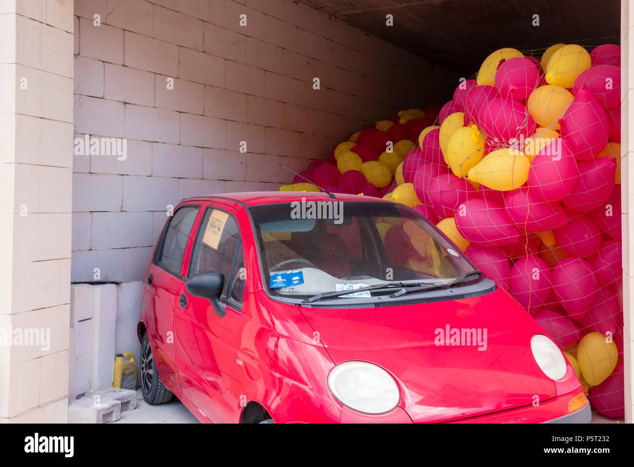 Red and yellow balloons trapped in a garage by a car in advance of a religious festival, Xwekija, Gozo, Malta Stock Photo