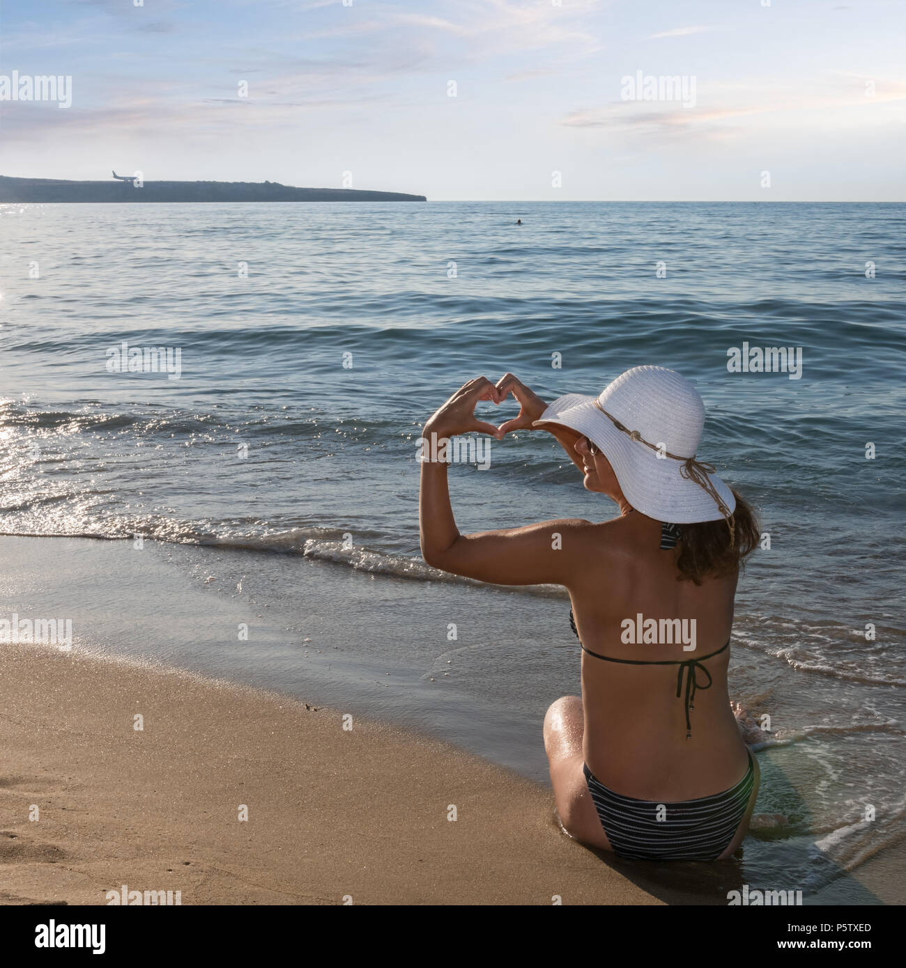 Woman with a sun hat on the seashore, forms with her fingers a heart shape Stock Photo