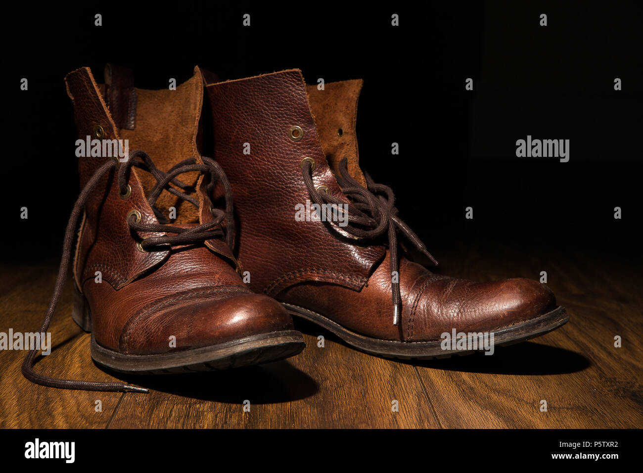Close up of men's traditional brown, leather ankle boots, laces undone, on wooden flooring with dark background. Artistic low-key lighting. Stock Photo