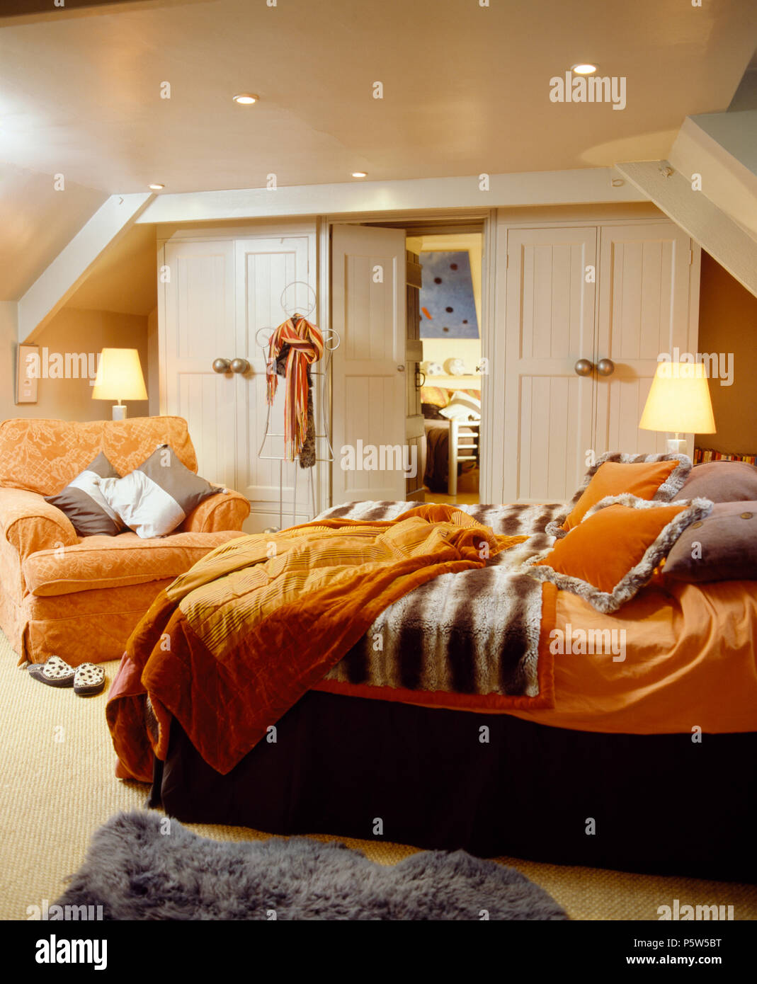 Orange quilt and linen on bed with faux fur throw on bed in attic bedroom with lighted lamps and orange armchair Stock Photo