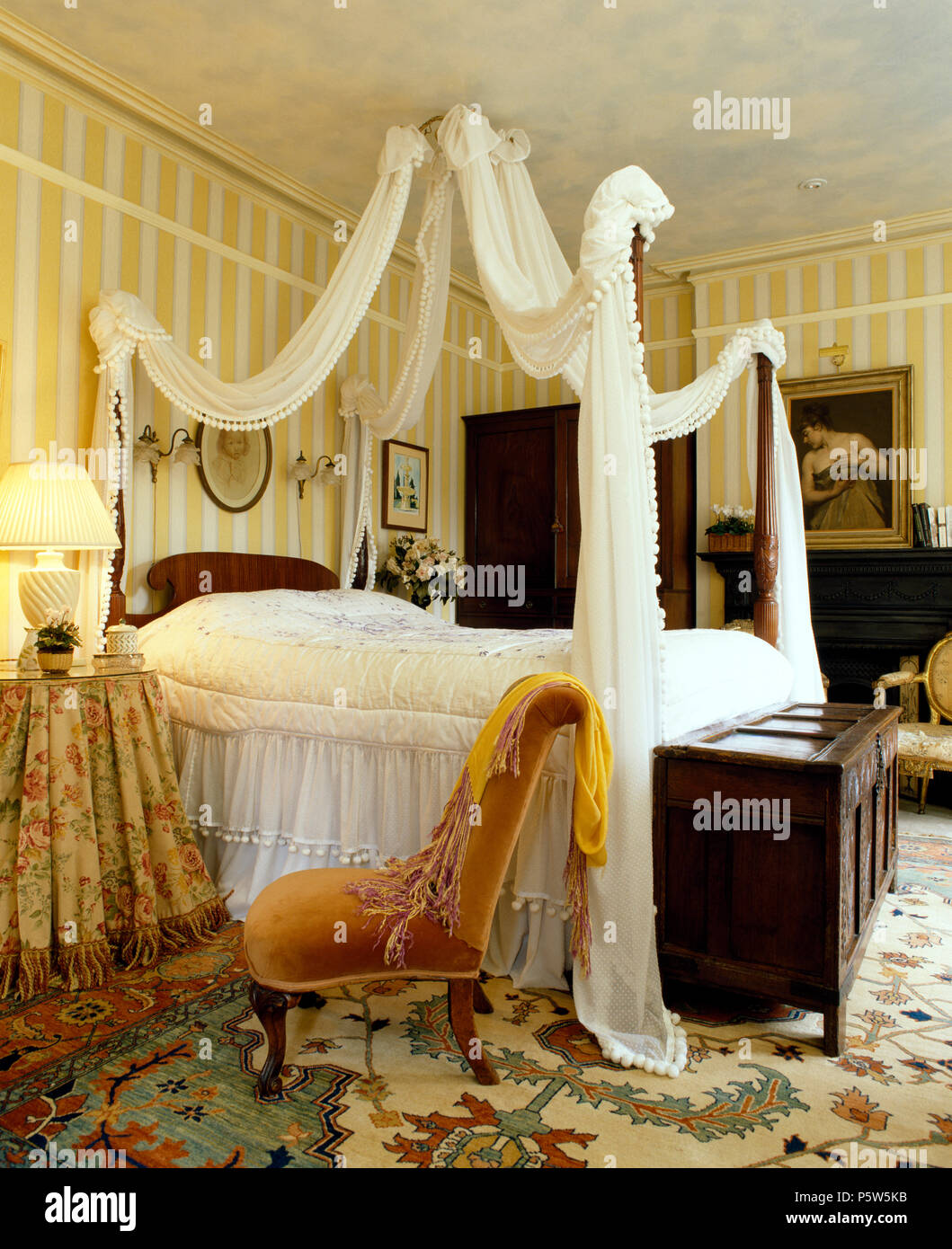 White voile drapes and valence on bed with spindle poles in bedroom with striped wallpaper and an antique chest Stock Photo
