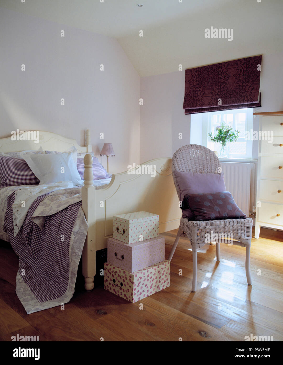 Painted vintage wicker chair beside cream painted bed with mauve quilt in attic bedroom with wooden flooring and purple blind Stock Photo