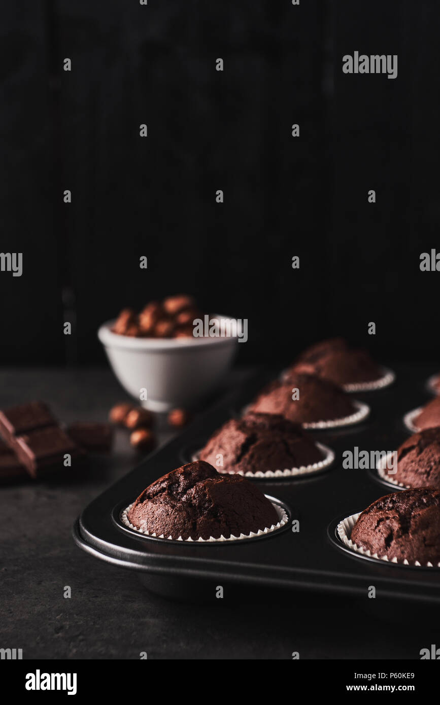 Chocolate muffins in bakeware with chocolate and nuts on the side on black concrete in front of a black wooden background with copy space. Stock Photo