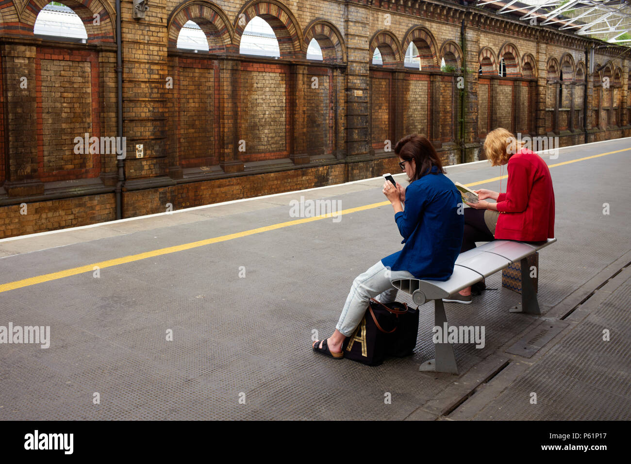 Two passengers waiting at Crewe station one reading a book and one looking at phone Stock Photo