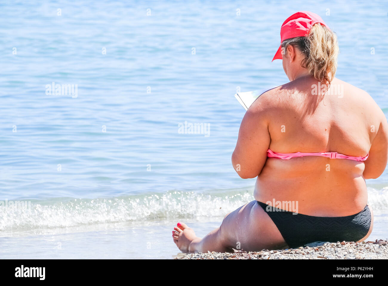 A woman wearing a hat, sitting on the beach, reading a book and staring at the sea.overweight female body on a beach.plus size woman. Copy space Stock Photo