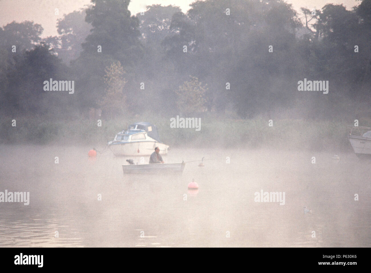Lone man fishing from small dinghy  at dawn, Oulton Broad, Suffolk, England, 1975 Stock Photo