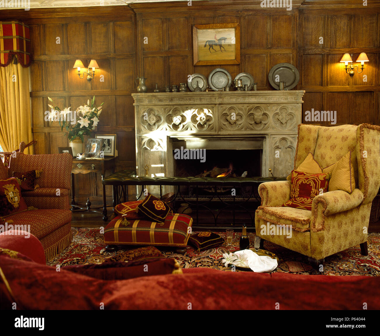 Upholstered stool and wing chair beside carved stone fireplace with club fender in panelled drawing room Stock Photo