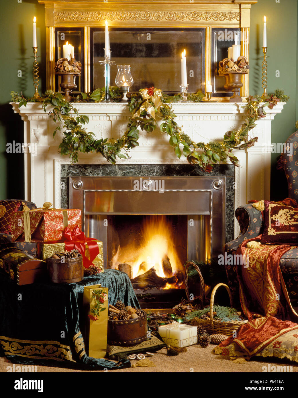 Fireplace with lighted firen and mantlepiece with lighted candles and foliage garland in sitting room with Christmas gifts piled on chair Stock Photo
