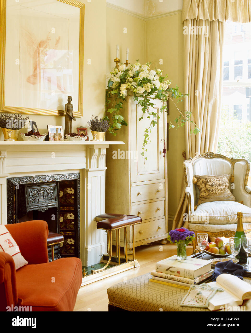 Club-fender on fireplace in cream living room with painted cupboard and cream armchair Stock Photo