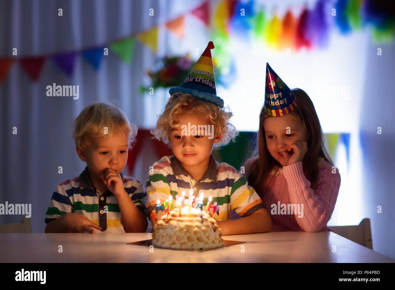 Kids birthday party. Children blow out candles on cake in dark room. Rainbow decoration and table setting for kids event, banner and flag. Girl and bo Stock Photo