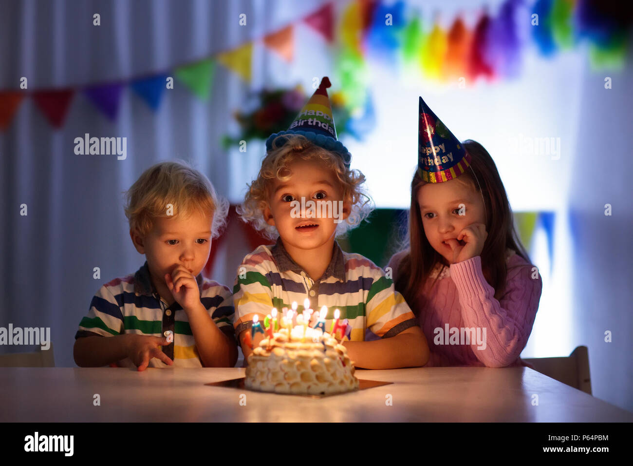 Kids birthday party. Children blow out candles on cake in dark room. Rainbow decoration and table setting for kids event, banner and flag. Girl and bo Stock Photo
