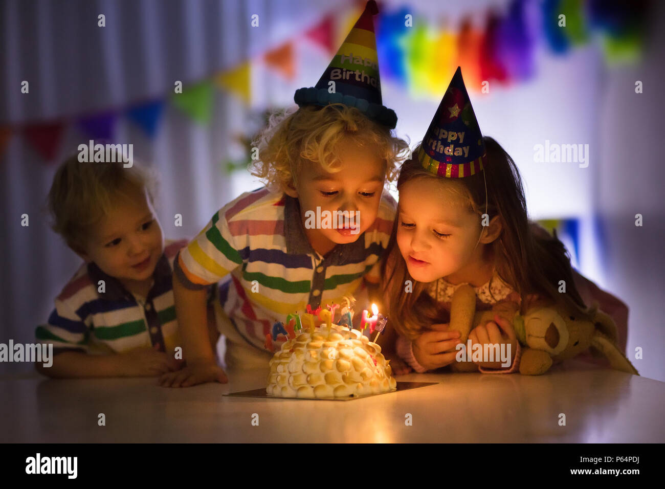 Kids birthday party. Children blow out candles on cake in dark room. Rainbow decoration and table setting for kids event, banner and flag. Girl and bo Stock Photo