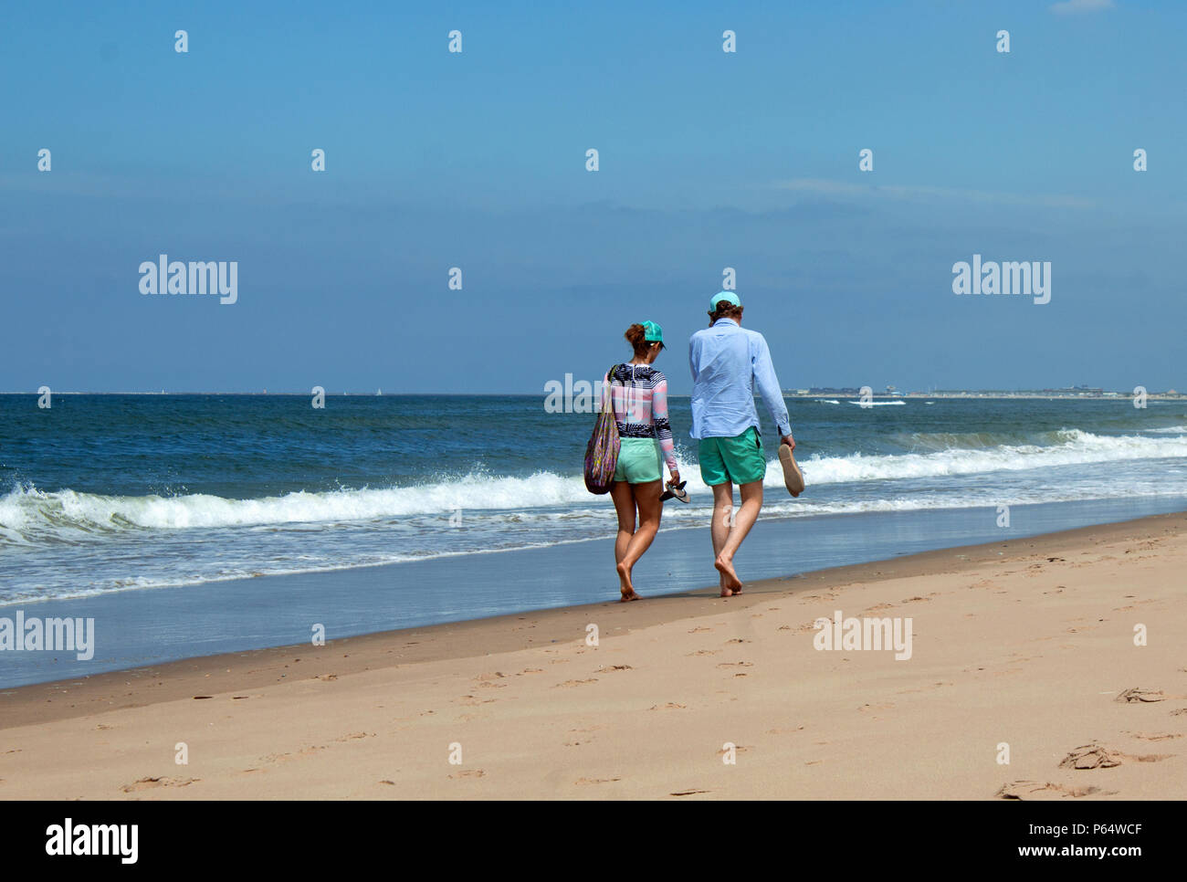 Ein junges, schlankes Paar von hinten, welches am Strand von Rügen spazieren geht. Beide tragen mit farbene Shorts und mit farbene Caps, beide barfuß. Stock Photo