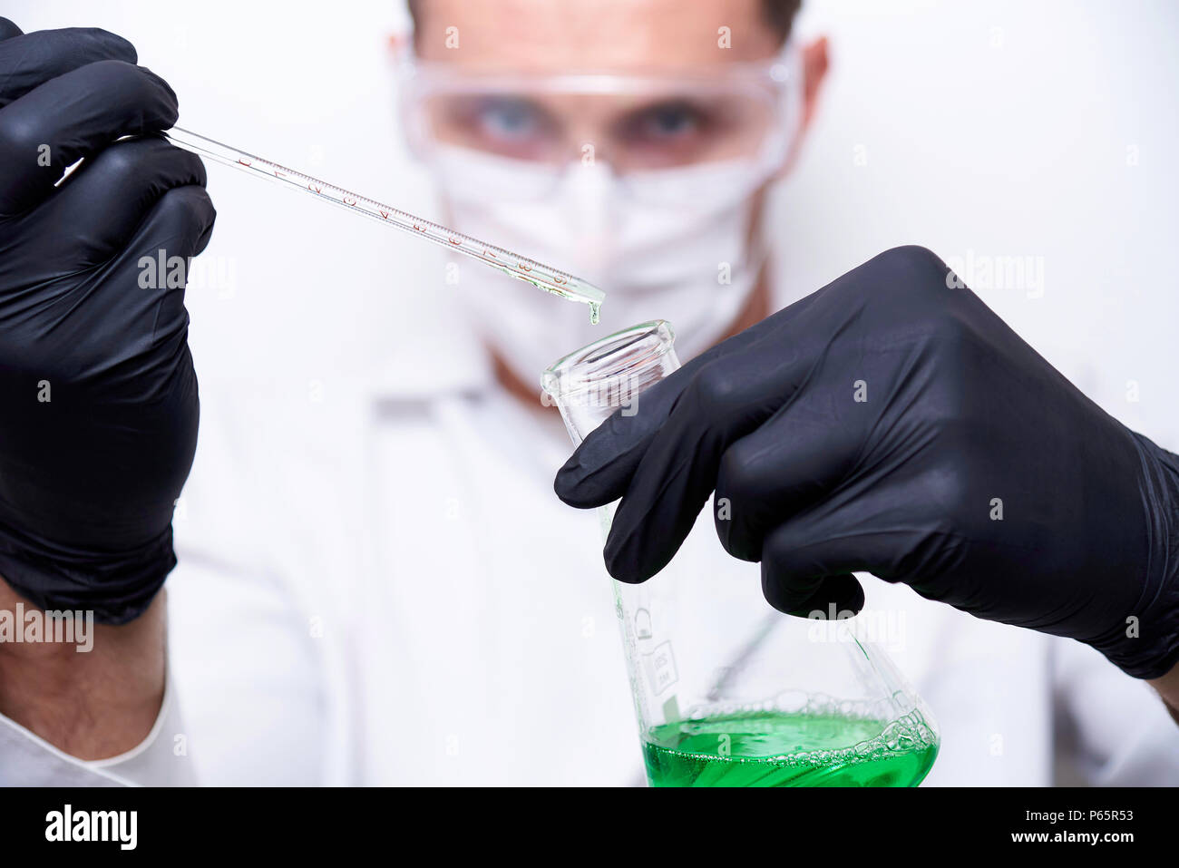 A test tube with a falling drop in a glass flask in the hands of a scientist wearing protective goggles, a mask and black gloves on a white background Stock Photo