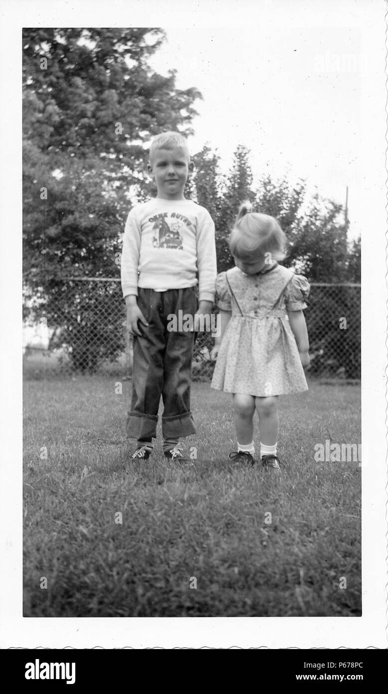 Black and white photograph, showing two small, light-haired children, standing together, facing the camera, in full length, the boy wears jeans turned up at the ankles and a long-sleeved shirt with a Gene Autry logo, the small girl wears a floral dress and looks down toward the grass at her feet, with a fence and foliage visible in the background, likely photographed in Ohio, 1955. () Stock Photo