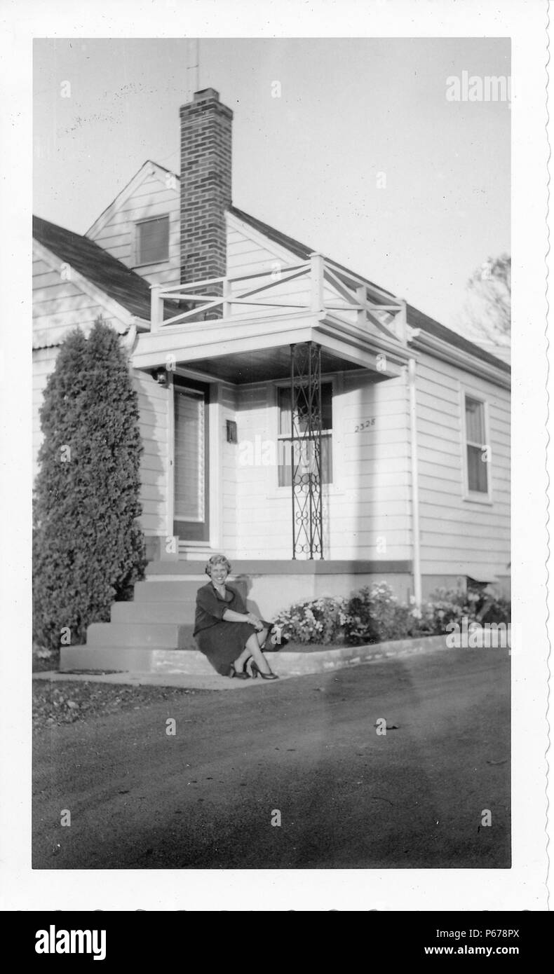 Black and white photograph, showing an attractive, smiling woman, with short, curly, blonde hair, wearing a skirt and heels, and sitting on the concrete border of a flower bed, in front of a small white house, likely photographed in Ohio in the decade following World War II, 1950. () Stock Photo