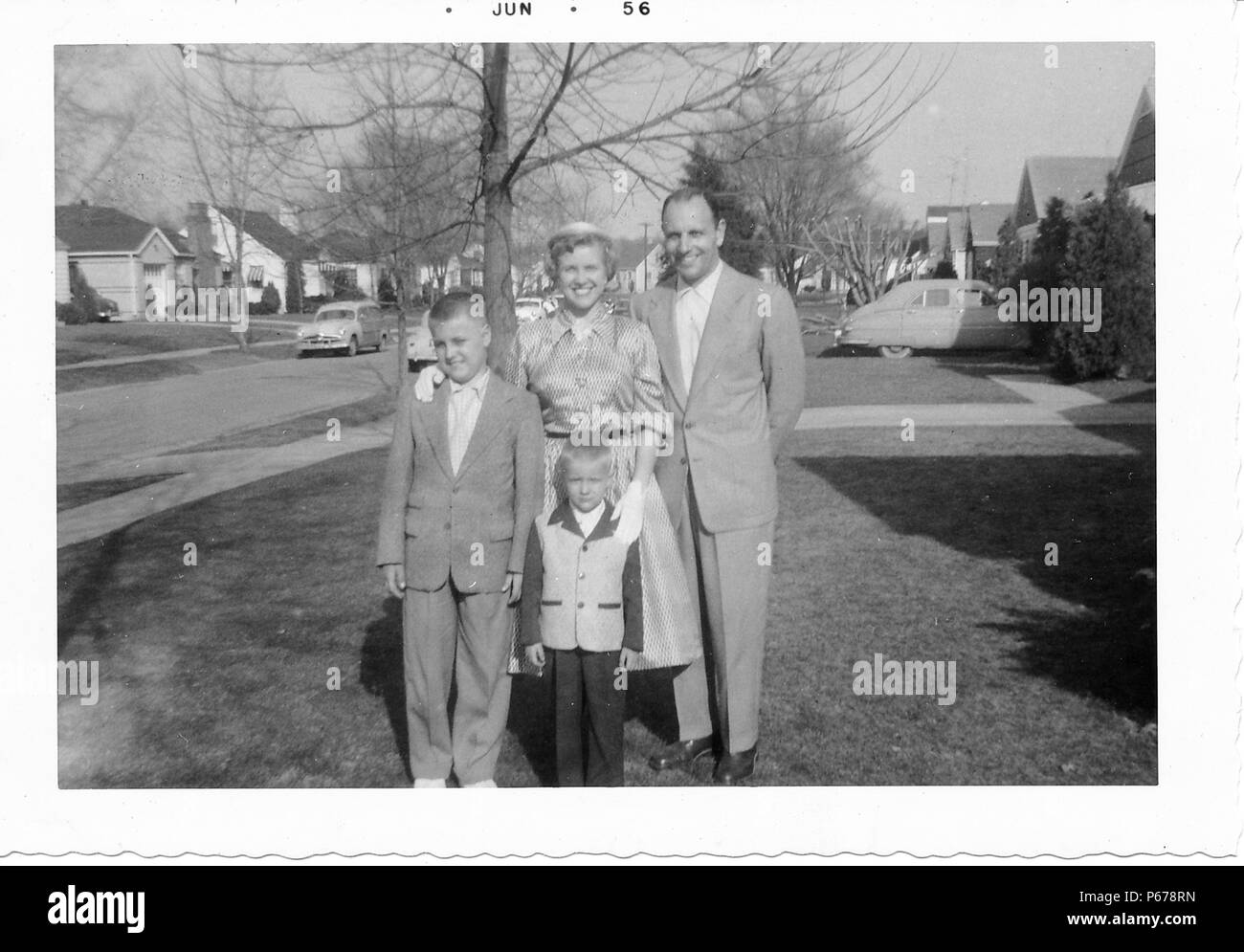 Black and white photograph, showing a family, in full length, posing together outdoors, the middle-aged man, with a receding hairline, wears a light colored suit and stands with a short-haired woman, wearing a skirt, blouse, and gloves, and two young boys, both wearing suits, the smaller one wearing a two-toned jacket, with trees, vintage cars, and suburban houses visible in the background, likely photographed in Ohio, June, 1956. () Stock Photo