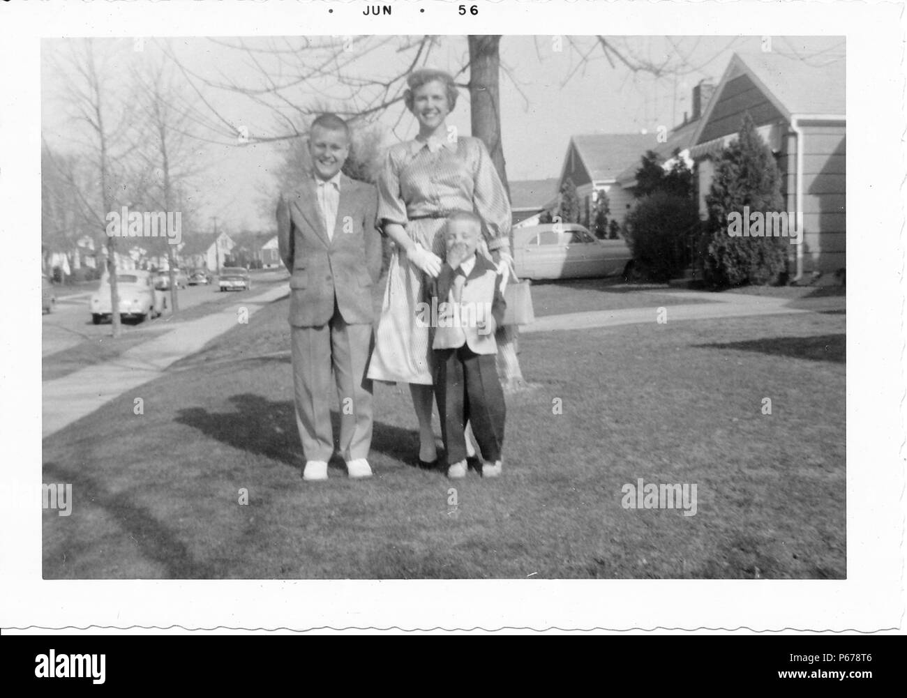 Black and white photograph, showing a short-haired woman, wearing a skirt, blouse, and gloves, posing with two young boys, both wearing suits, the smaller one wearing a two-toned jacket, with trees, vintage cars, and suburban houses visible in the background, likely photographed in Ohio, June, 1956. () Stock Photo