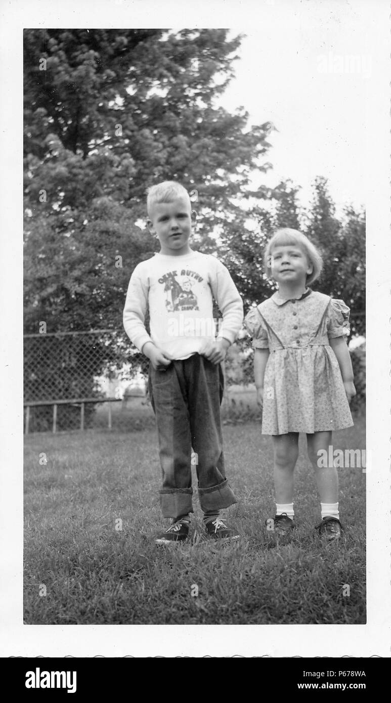 Black and white photograph, showing two small, light-haired children, standing together, facing the camera in full length, the boy wears jeans turned up at the ankles and pulls at the hem of his long-sleeved shirt to emphasize the Gene Autry logo, the small girl wears a floral dress and makes a face at the camera, with a fence and foliage visible in the background, likely photographed in Ohio, 1955. () Stock Photo