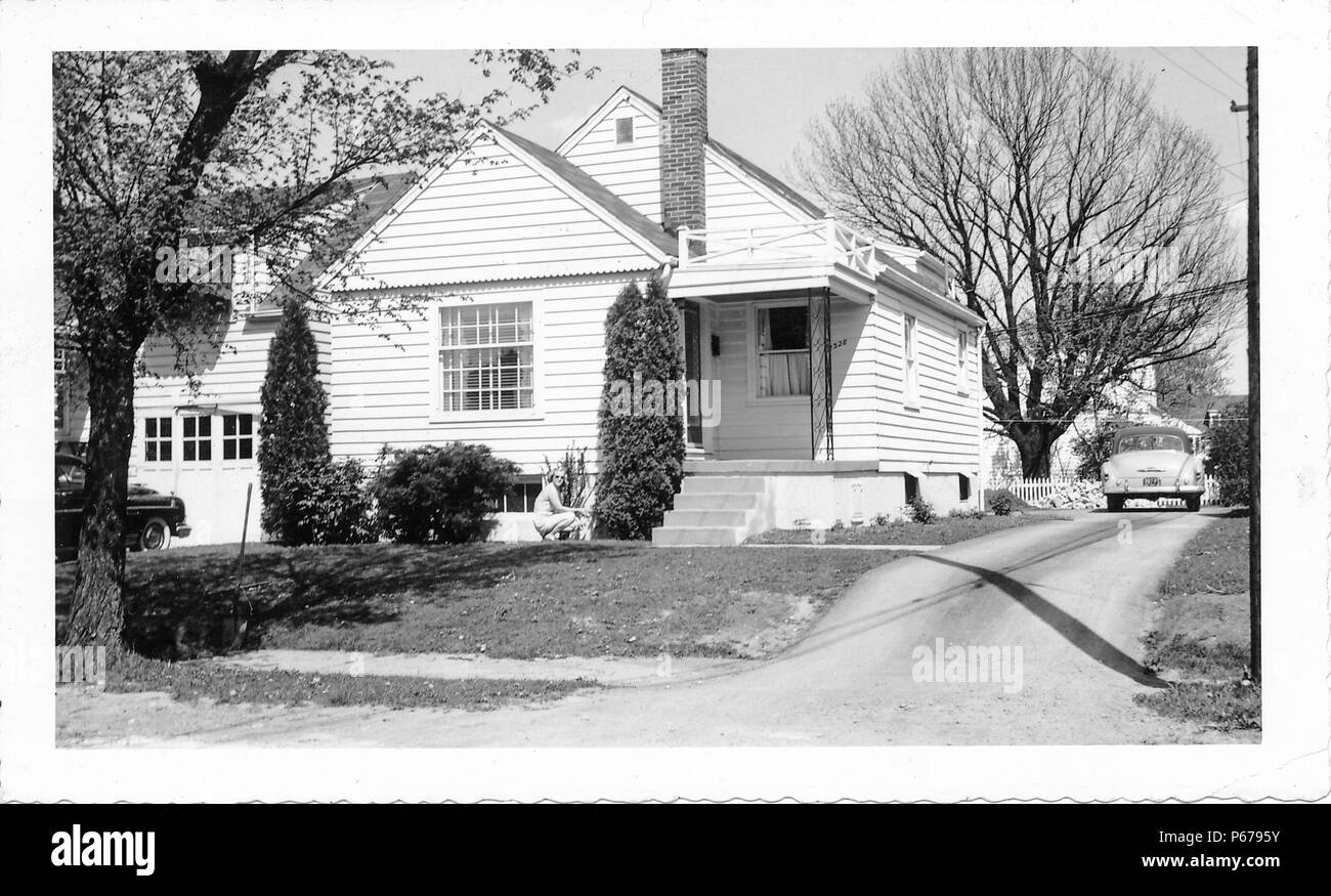 Black and white photograph, showing a blonde woman, wearing sunglasses, and crouching down (presumably to garden) in front of a small, light-colored, tract house, with siding, a covered front porch, and a vintage car at the end of the driveway, likely photographed in Ohio in the decade following World War II, 1950. () Stock Photo