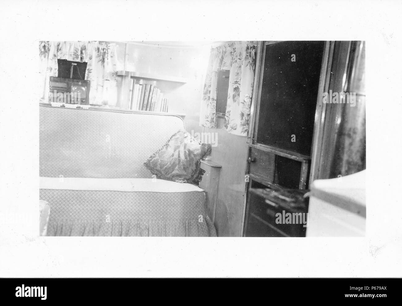 Black and white photograph, showing the cramped interior of a Mid-century caravan or trailer home, with a sofa, bookshelves, a vintage radio, and a door visible at right, likely photographed in Ohio in the decade following World War II, 1950. () Stock Photo