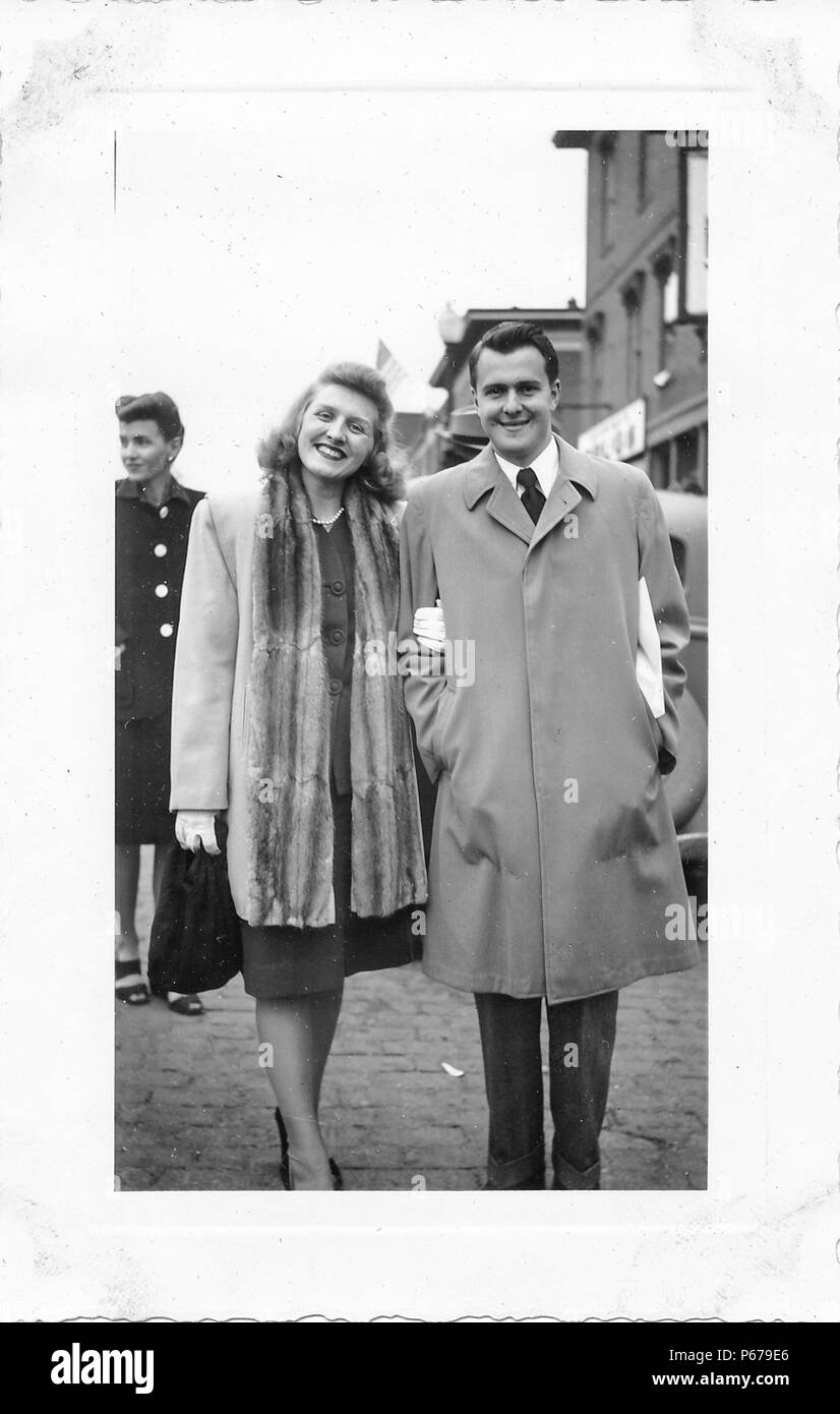 Black and white photograph, showing a well-groomed, smiling man and woman, in full length, facing the camera, the man wearing a light-colored, single-breasted trench coat, and the woman wearing a mid-length coat, with a long fur, stole or lapel, over a two-piece skirt suit, and a string of pearls around her neck, with a second, carefully-coiffed, dark-haired woman in a skirt suit and heeled sandals visible in the background, likely photographed in Ohio in the decade following World War II, 1950. () Stock Photo