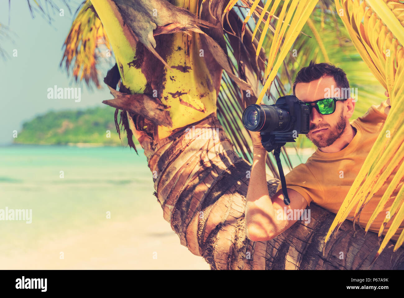 Male Paparazzi hides behind a tree on a tropical beach to take pictures of a hidden camera Stock Photo