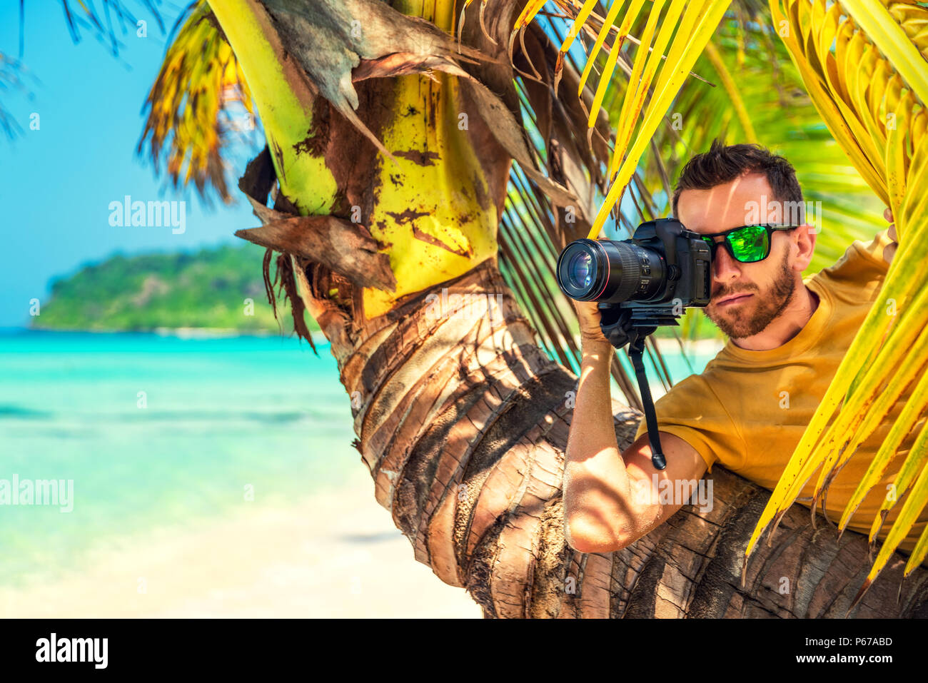Male Paparazzi hides behind a tree on a tropical beach to take pictures of a hidden camera Stock Photo
