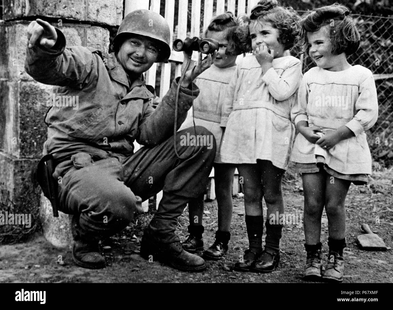 Image shows an American soldier holding binoculars up to three little girls so they're able to glance through the lens. After the liberation of Normandy, during World War 2 1944. Stock Photo