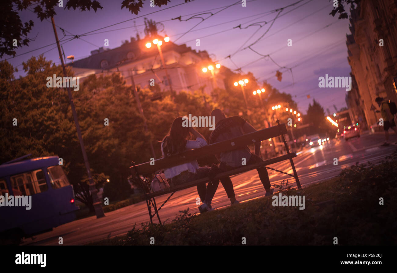 Rear view of a young couple sitting on a bench by the street, enjoying a warm summer night in Zagreb, Croatia. Stock Photo