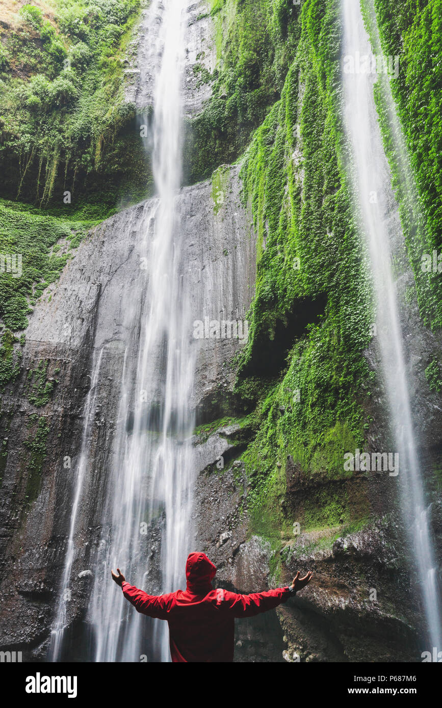 Traveller in red jacket raised hand, at Madakaripura waterfall in Indonesia. Travel lifestyle and success Stock Photo