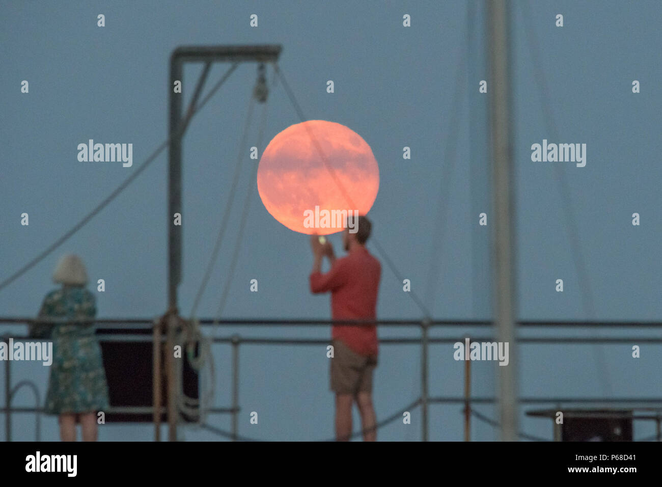 Mousehole, Cornwall, UK. 28th June 2018. UK Weather. A group of friends stand on the harbour wall at Mousehole, watching the full Strawberry moon rise. Credit: cwallpix/Alamy Live News Stock Photo