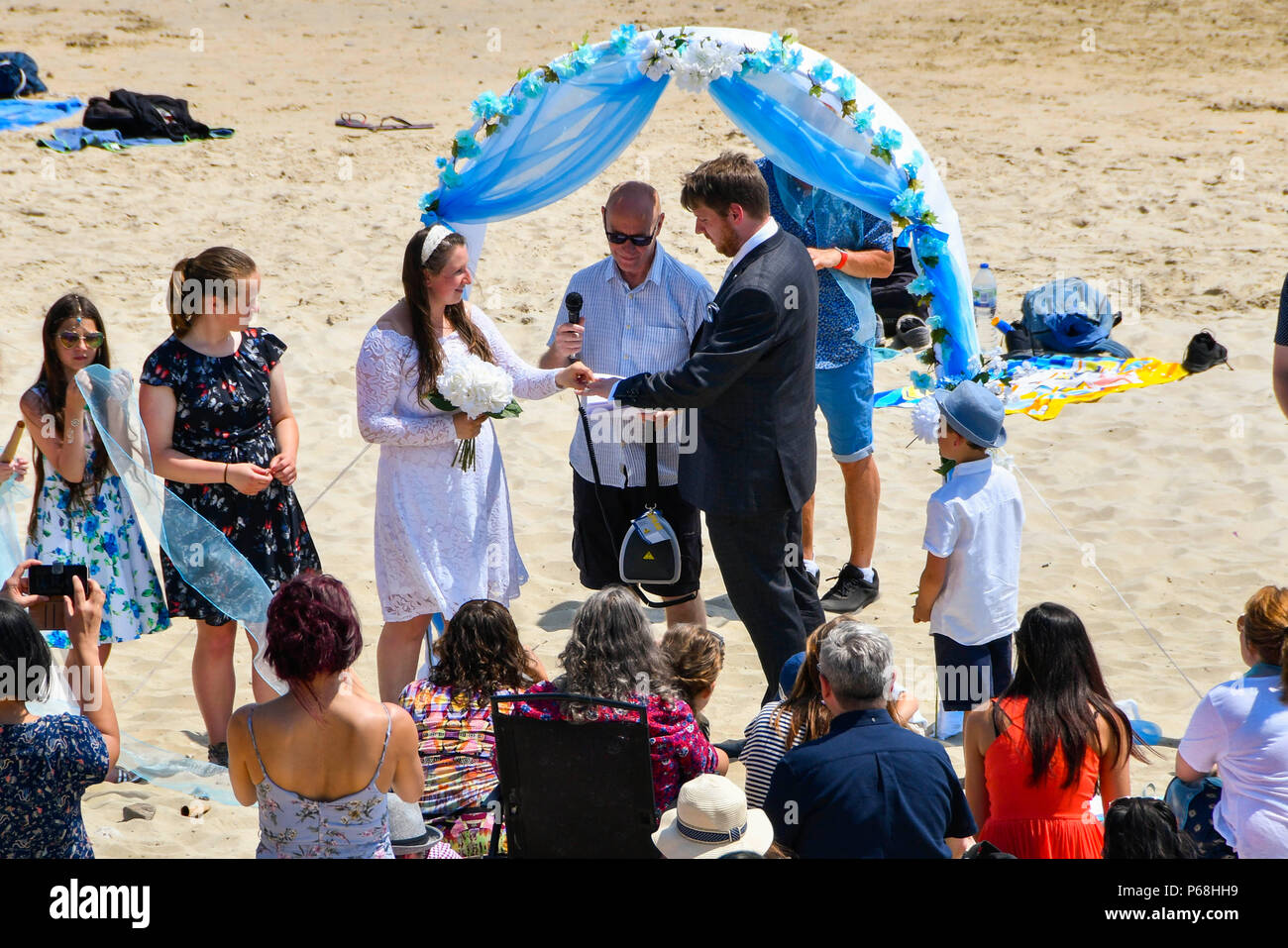 Lyme Regis, Dorset, UK.  29th June 2018. UK Weather.  A couple, Kashi and Michael Morrish having their wedding on the beach surrounded by guests and sunbathers at the seaside resort of Lyme Regis in Dorset on a day hot sunshine and clear blue skies as the heatwave continues.  Kashi puts the ring on Michael's finger.  Picture Credit: Graham Hunt/Alamy Live News Stock Photo