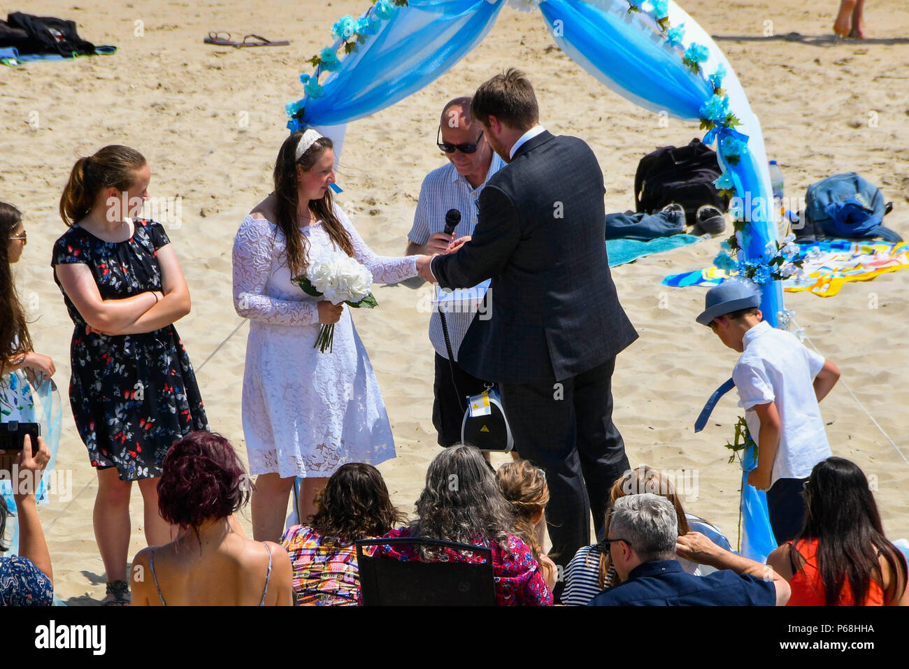 Lyme Regis, Dorset, UK.  29th June 2018. UK Weather.  A couple, Kashi and Michael Morrish having their wedding on the beach surrounded by guests and sunbathers at the seaside resort of Lyme Regis in Dorset on a day hot sunshine and clear blue skies as the heatwave continues.  Micheal puts the ring on Kashi's finger.  Picture Credit: Graham Hunt/Alamy Live News Stock Photo