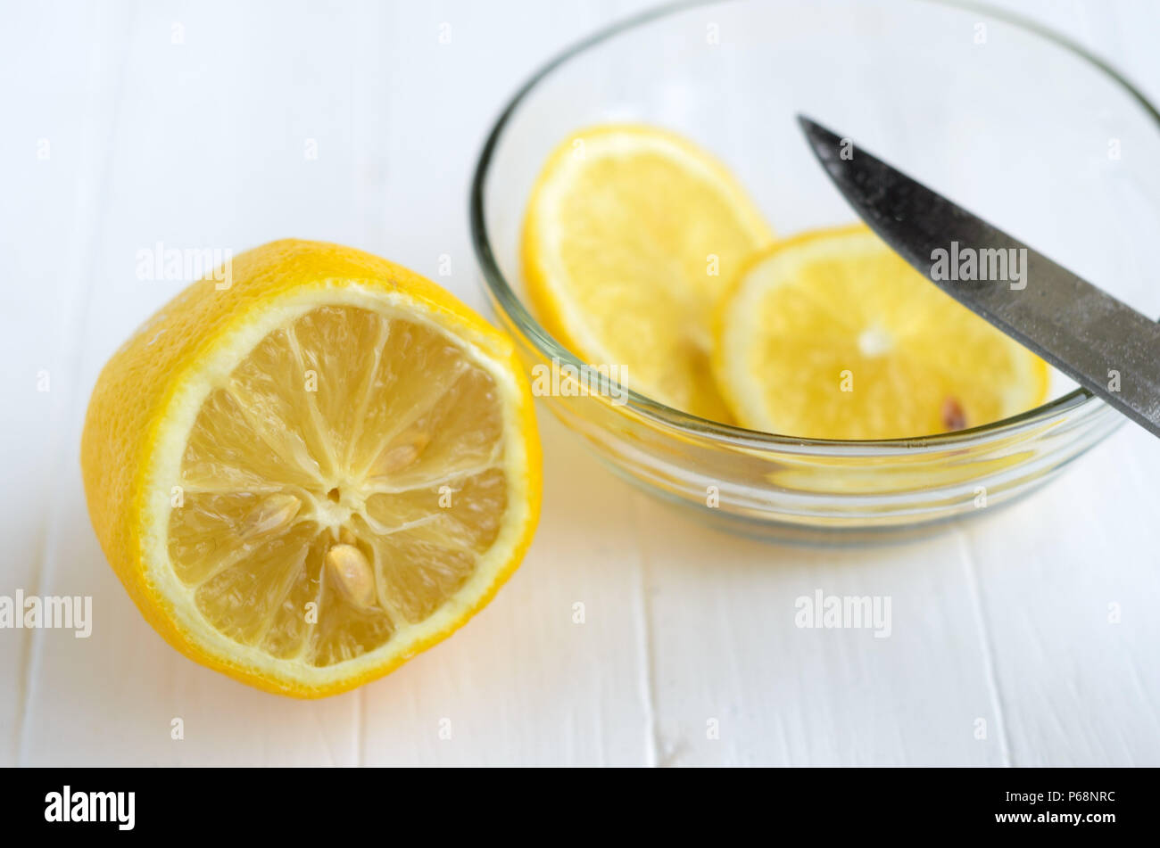 One cutted lemon and two slices of lemon into glass plate.Part of the knife blade on the glass plate. White wooden background. Stock Photo
