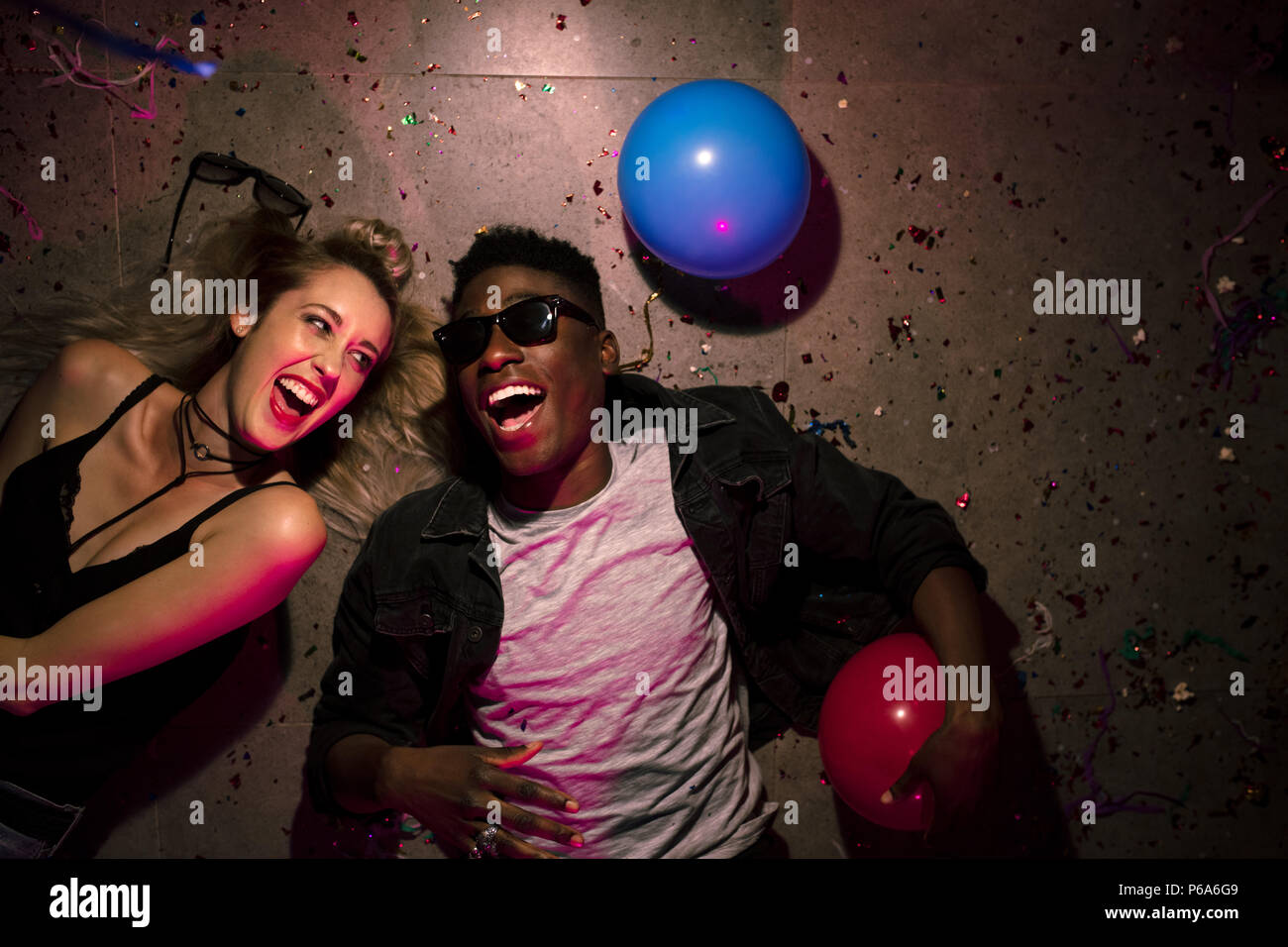 Top view of man and woman lying on floor and laughing at a house party. Friends lying on floor at the house party with balloons and confetti around. Stock Photo