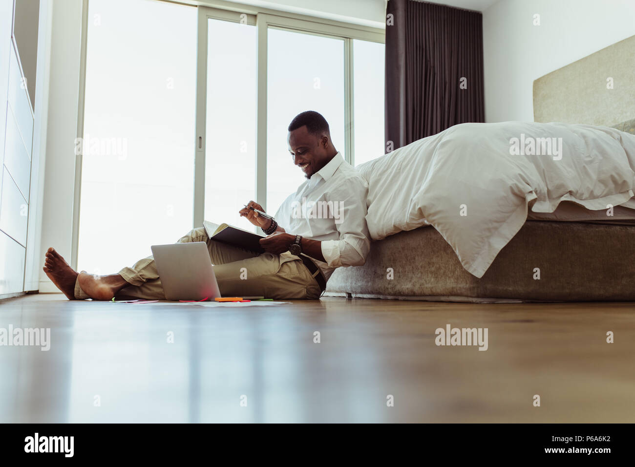 businessman reading his diary and smiling while working on laptop computer at home. Man working on laptop computer sitting on the floor. Stock Photo
