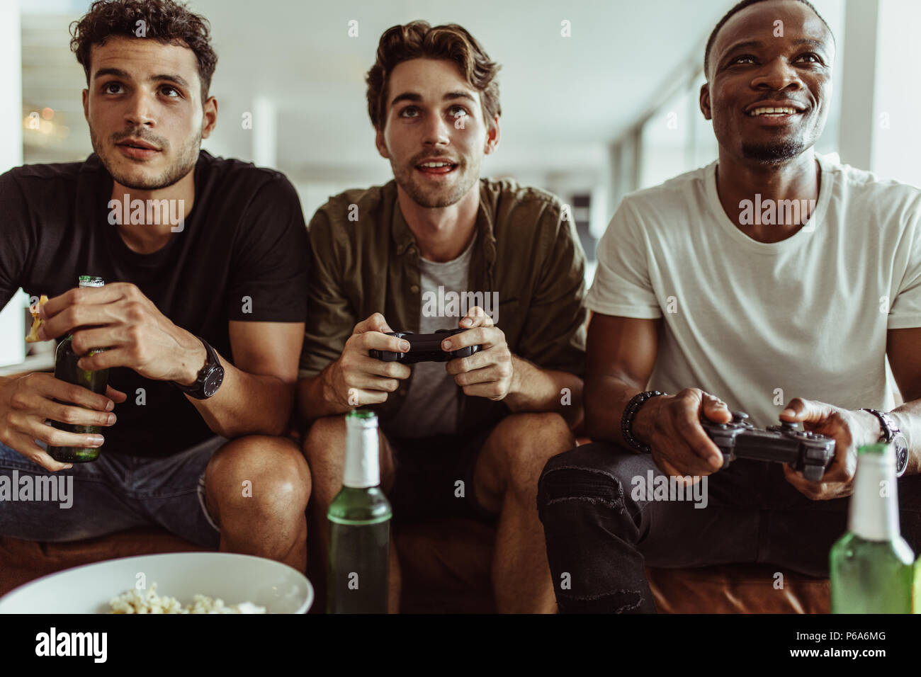 Friends having fun with drinks and snacks while playing video game at home. Two men playing video game holding joysticks while another man watches wit Stock Photo
