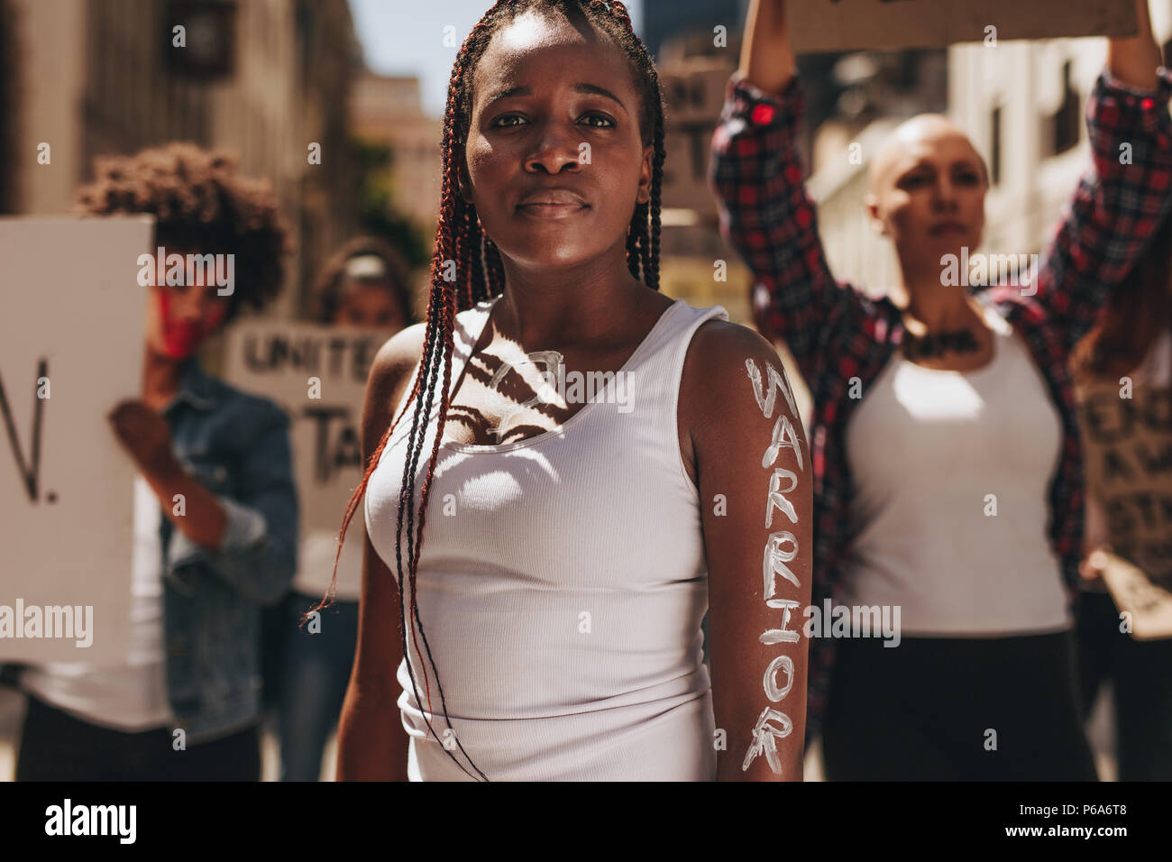 Young woman standing outdoors with group of protesters at back. Woman with word warrior written on her arm. Stock Photo