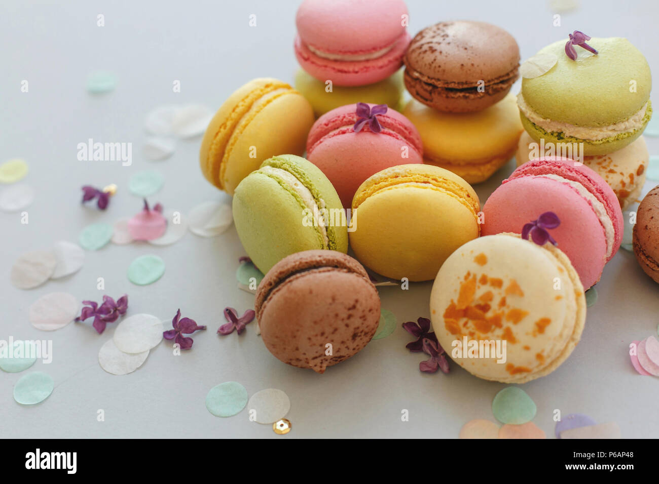 colorful macarons on trendy pastel gray paper with lilac flowers and confetti. tasty pink, yellow, green and brown macaroons. candy bar for party. foo Stock Photo