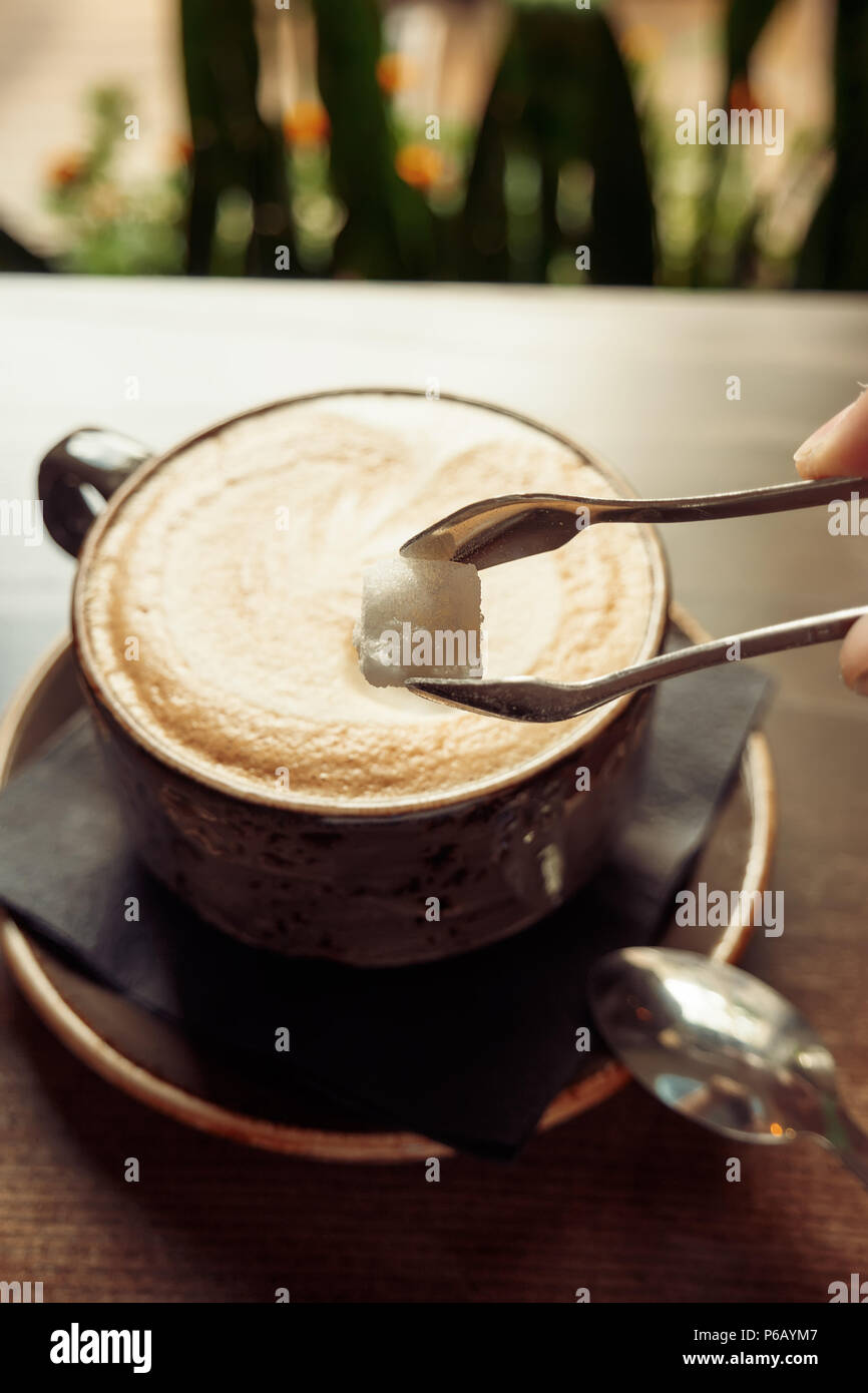 Close-up of adding cube of sugar into coffee cup on wooden table. Stock Photo