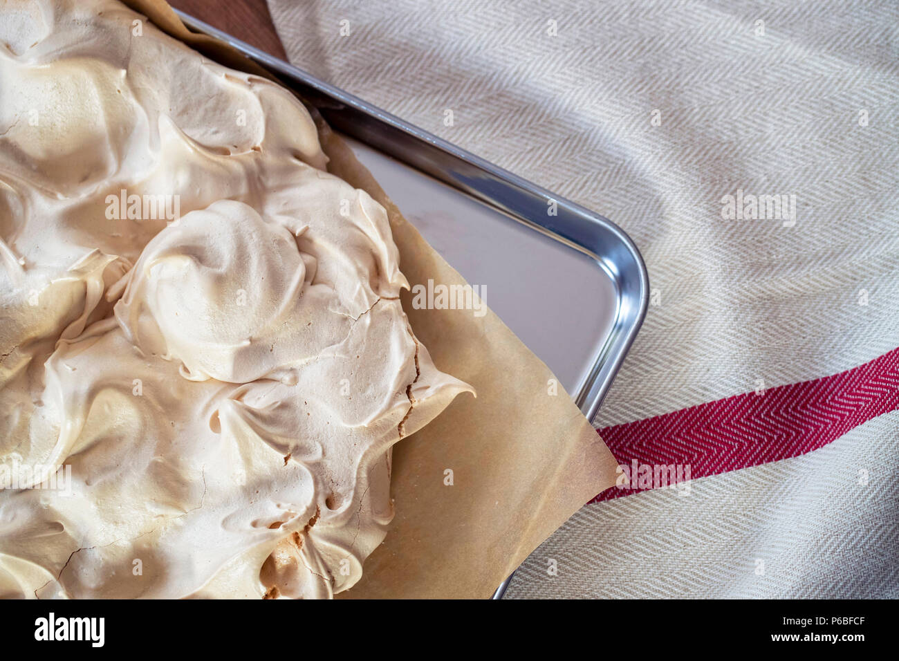 Cooking the meringue base for a Pavlova dessert. Fresh meringue, with crisp golden top, on baking parchment and a stainless steel baking tray. Overhea Stock Photo