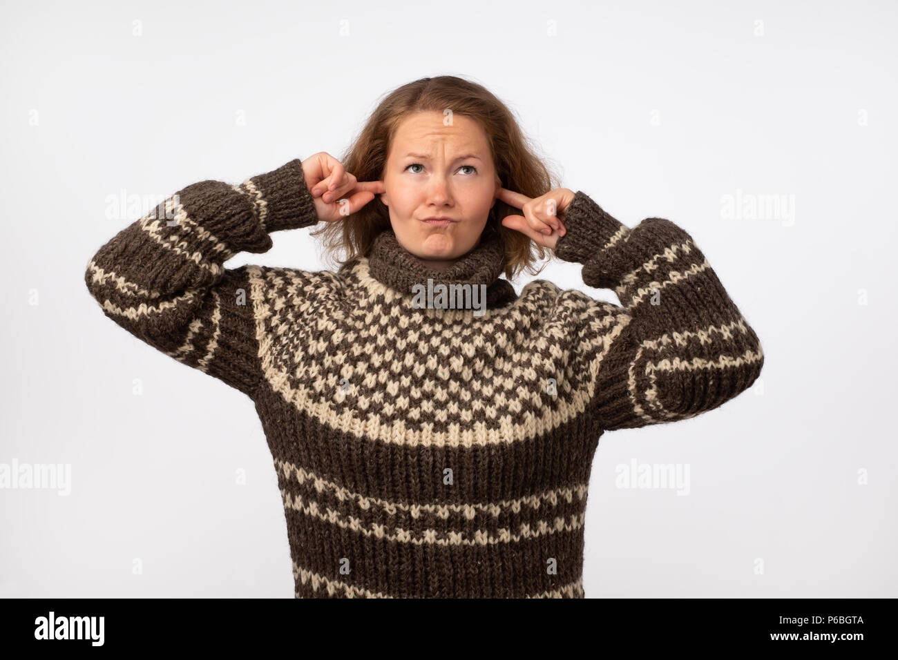 Young female in warm brown sweater plugging her ears and frowning her face being annoyed with noise. She wants to stay in calm comfortable place being Stock Photo