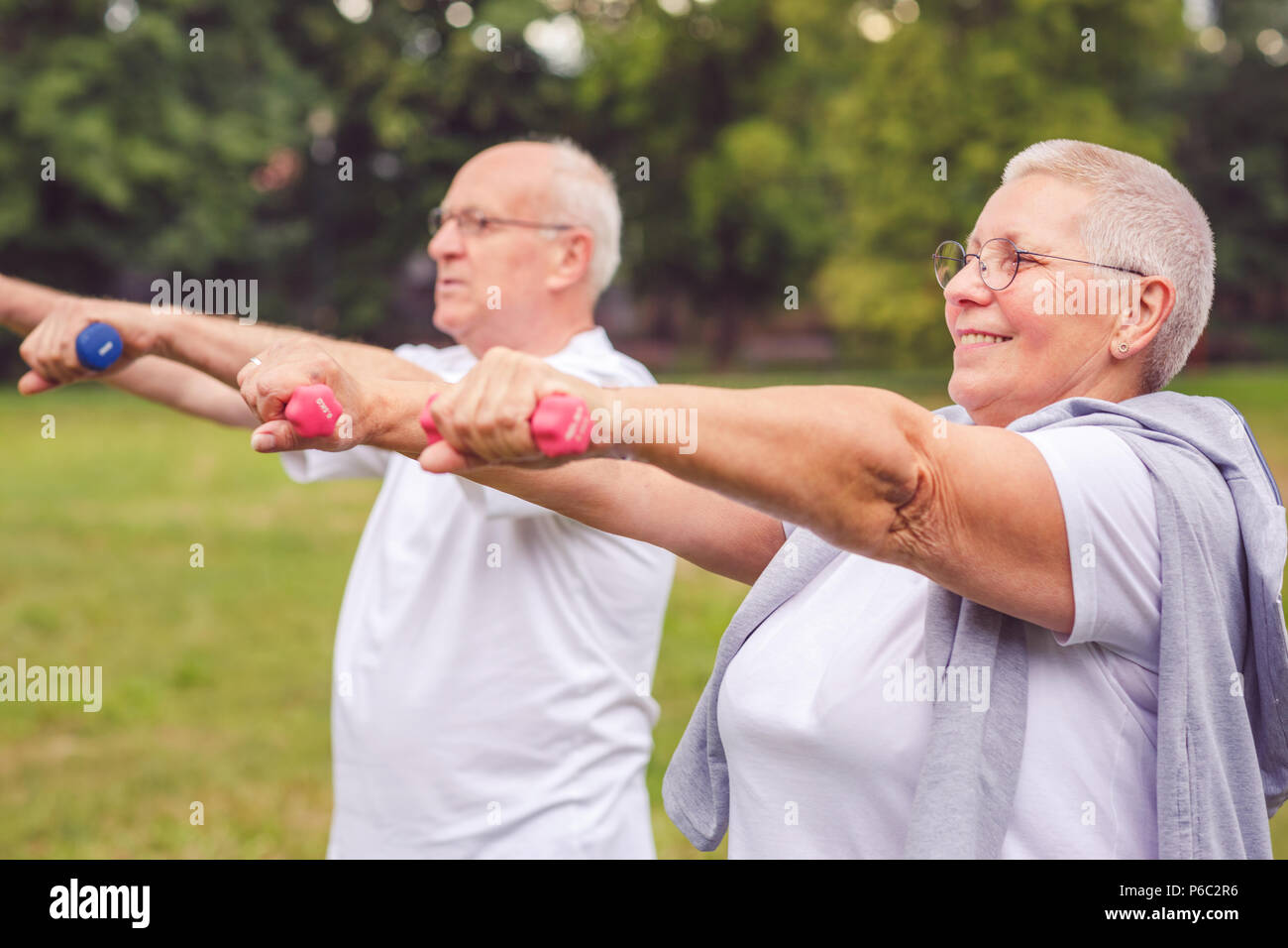 Smiling senior man and woman practice fitness exercise with dumbbells in a park at sunny day Stock Photo